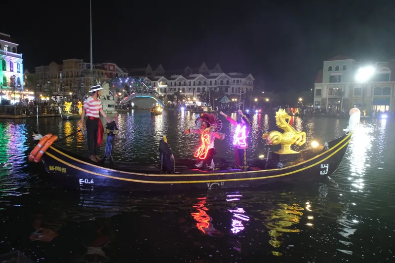 Performers in glowing LED costumes dance on a gondola at a night parade on the canal in Grand World, Phu Quoc. Performers in glowing LED costumes dance on a gondola at a night parade on the canal in Grand World, Phu Quoc.