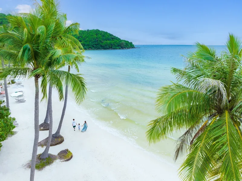 A family walks along a white sand beach fringed by palm trees with turquoise water in Phu Quoc.