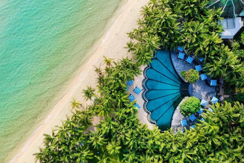 Aerial view of a fan-shaped resort pool nestled among palm trees beside a white sand beach in Phu Quoc. Aerial view of a fan-shaped resort pool nestled among palm trees beside a white sand beach in Phu Quoc.
