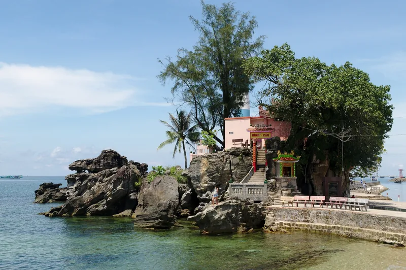 Dinh Cau temple sits atop rocky outcrops by the sea in Phu Quoc, shaded by a large tree. Dinh Cau temple sits atop rocky outcrops by the sea in Phu Quoc, shaded by a large tree.
