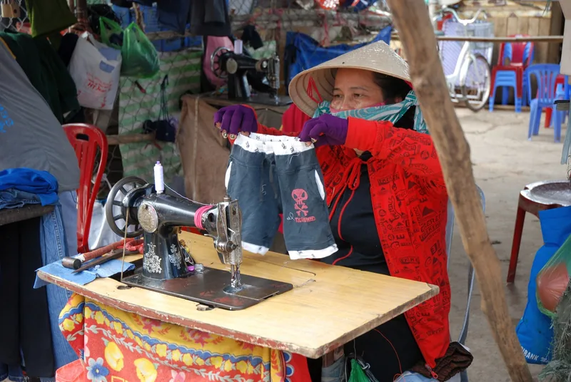 A woman wearing a conical hat sews clothing on a manual sewing machine at a market stall in Phu Quoc.