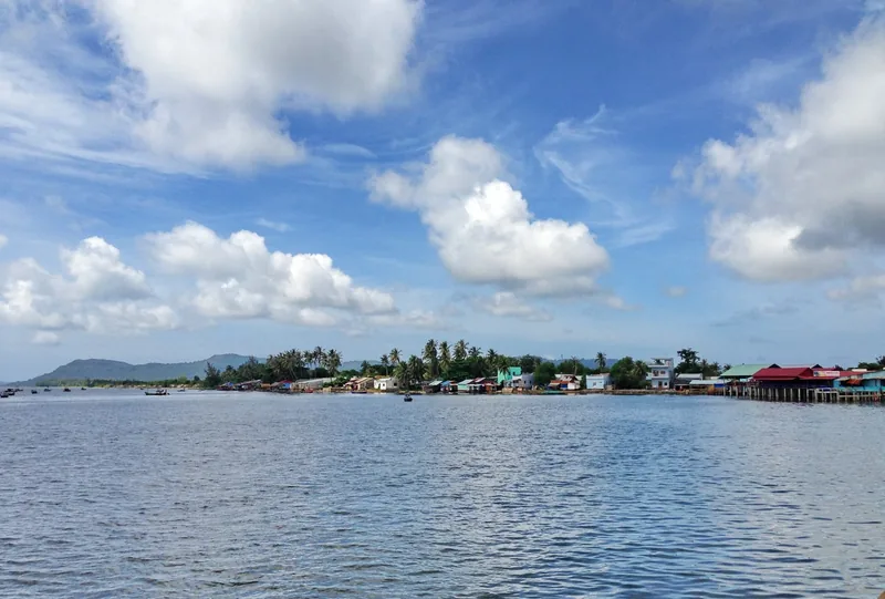 A coastal fishing village with colorful stilt houses and palm trees lines the shore of Phu Quoc under a broad blue sky.