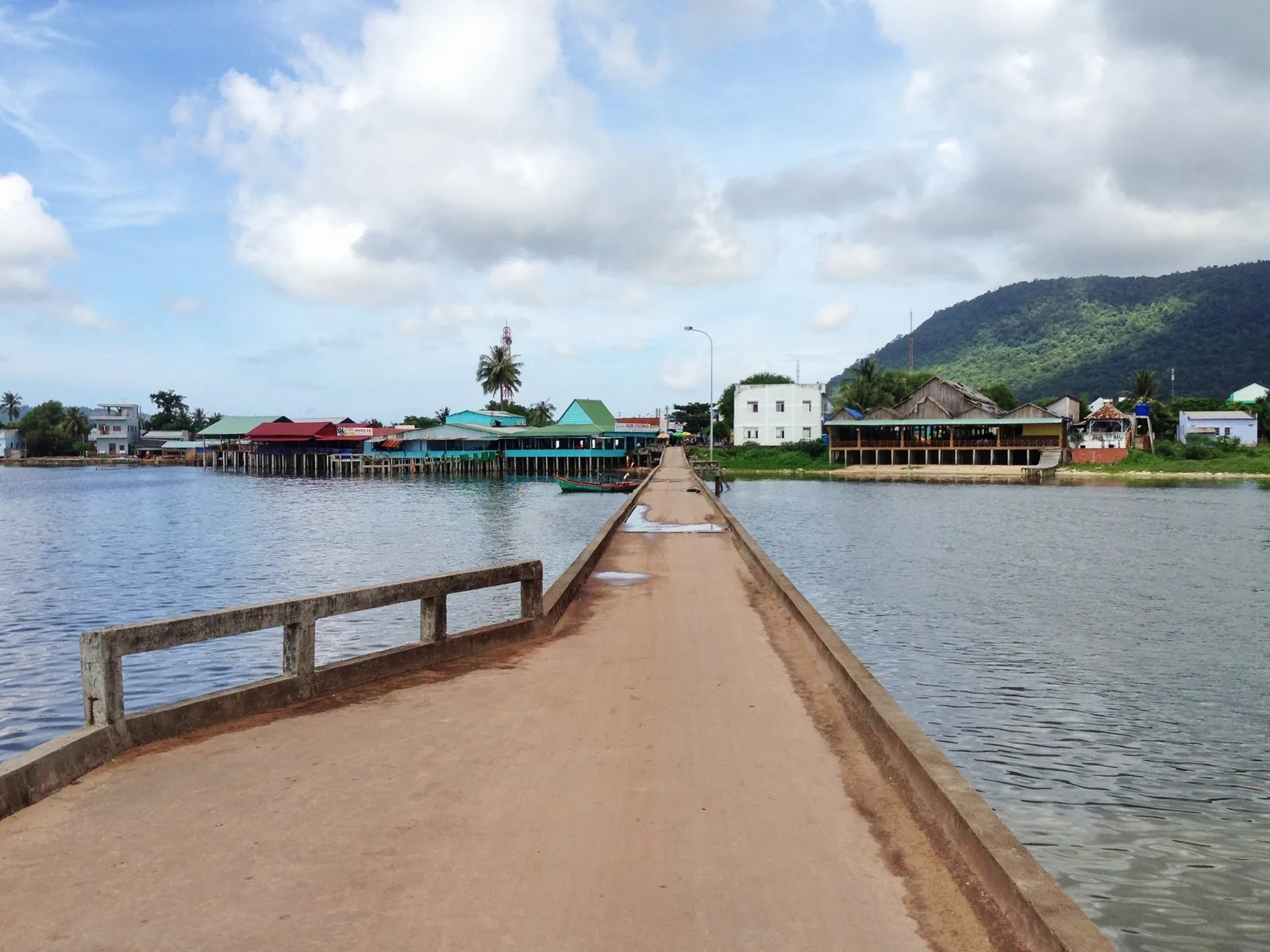 A concrete pier stretches over calm water toward a coastal village with colorful stilt houses on Phu Quoc island.