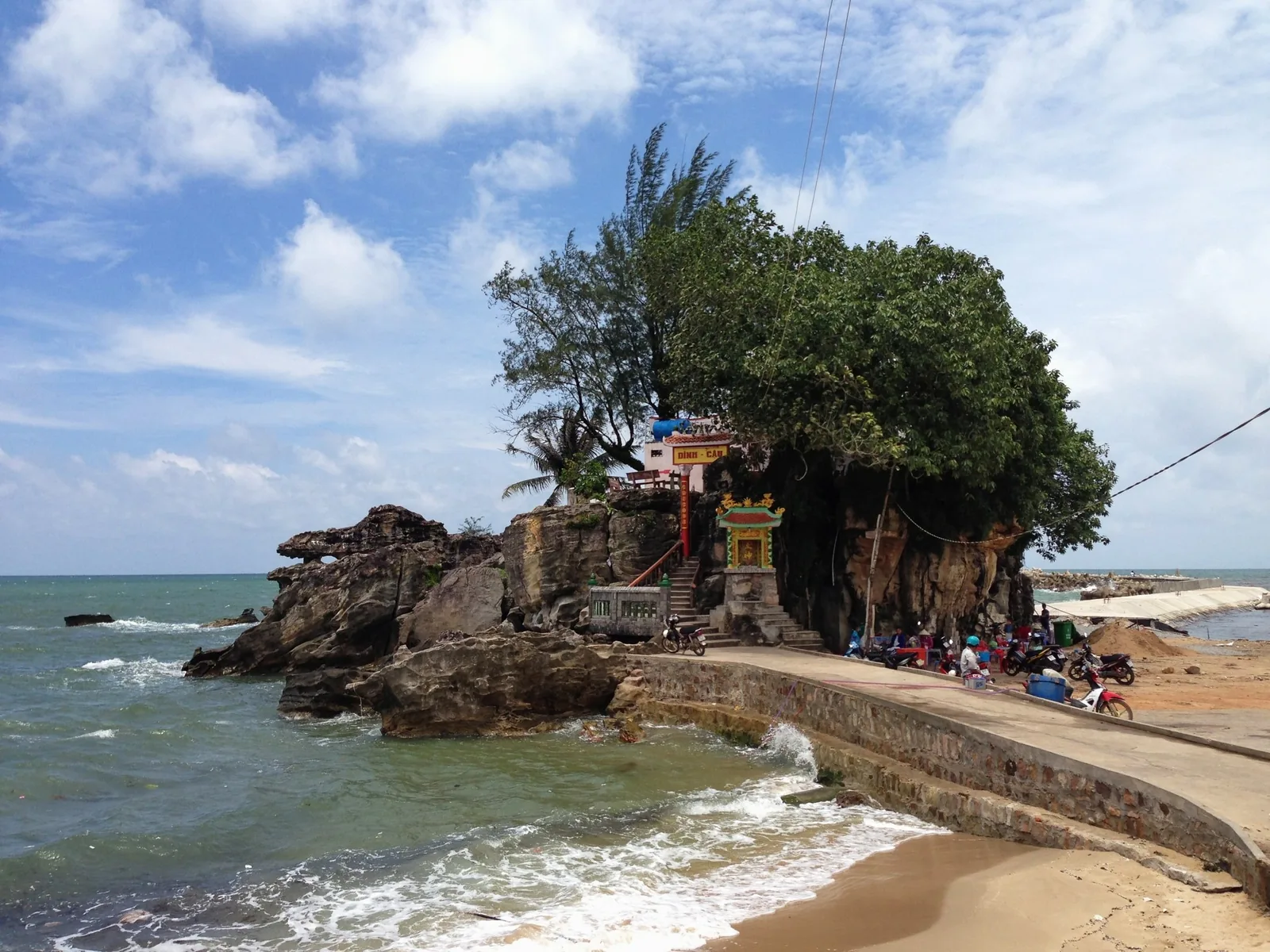 A small shrine perched on a rocky outcrop with a large tree at Dinh Cau in Phu Quoc, waves lapping the coastal path below.