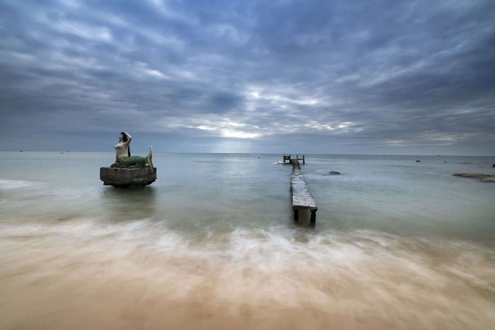 A mermaid statue on a rock formation stands beside a weathered wooden pier on Phu Quoc's beach under cloudy skies. A mermaid statue on a rock formation stands beside a weathered wooden pier on Phu Quoc's beach under cloudy skies.
