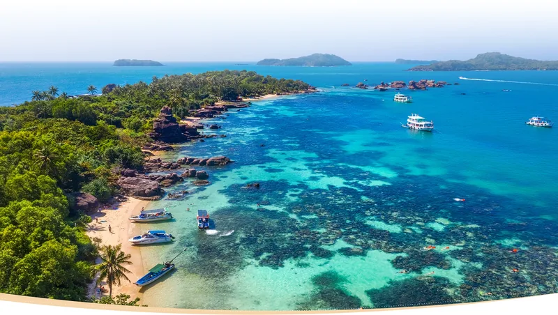 Aerial view of turquoise water, coral reef and boats along a lush coastline in Phu Quoc