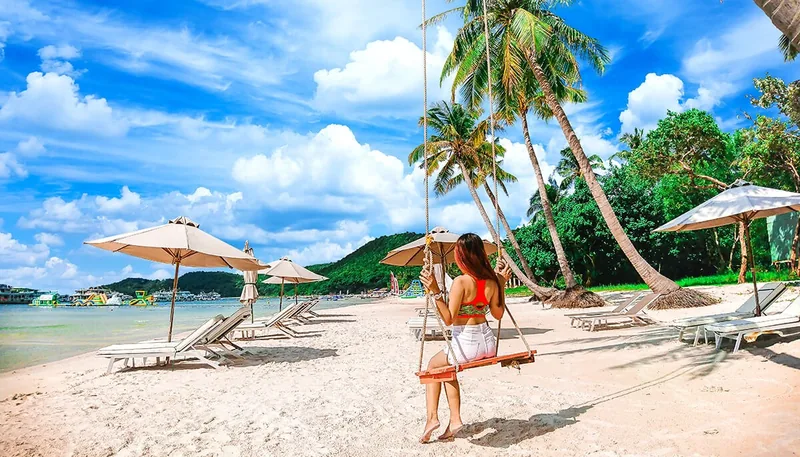 A woman swings on a beach in Phu Quoc, surrounded by palm trees, sun loungers and turquoise sea.