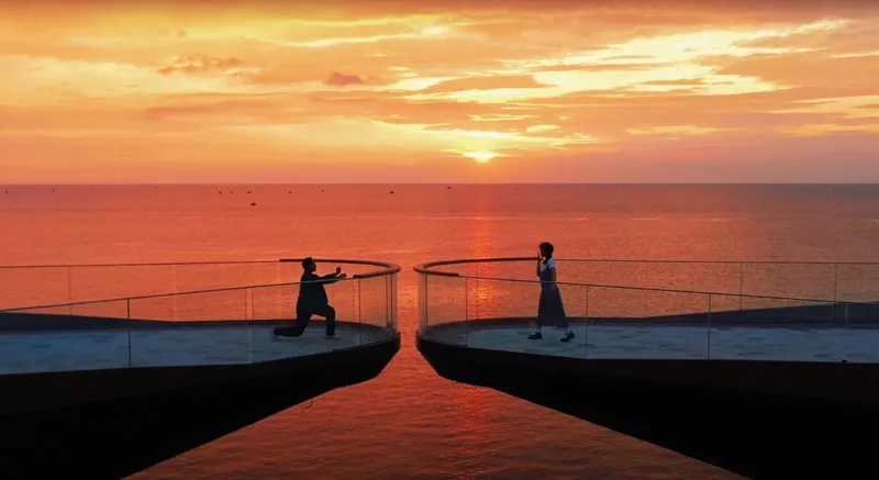 Two people stand on curved twin viewing platforms over the sea in Phu Quoc during a vivid orange sunset.